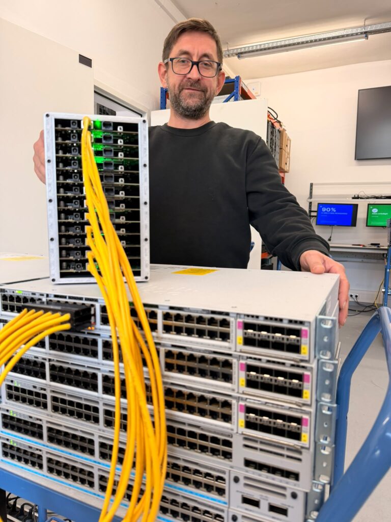 Testplatz mit gestapelten Cisco Catalyst Switches, Prüfgerät und gelben Patchkabeln, dahinter eine Person